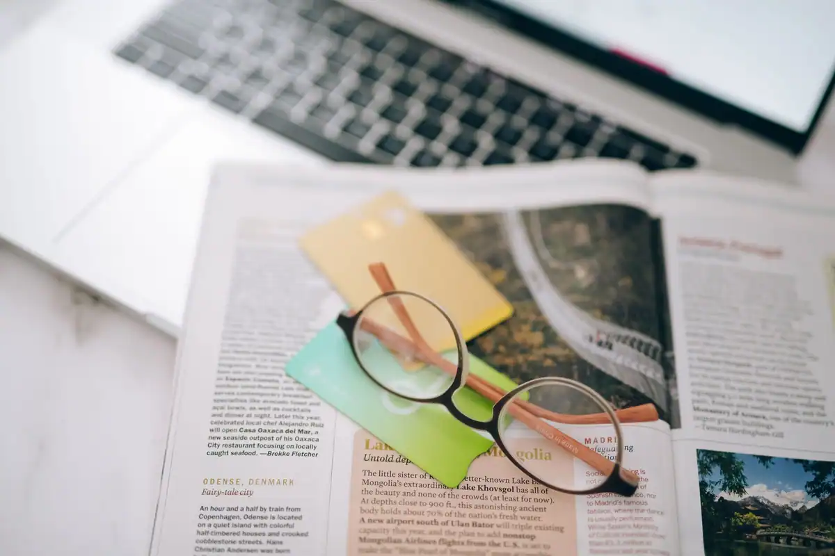 Ochelari de citit, carduri bancare și o revistă de călătorii lângă laptop